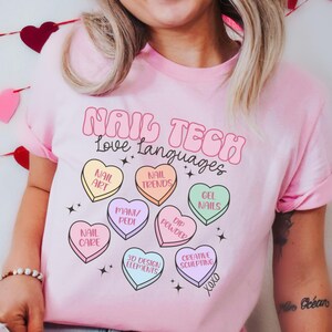 May include: A light pink t-shirt with the text "NAIL TECH Love Languages" in a bubble font. The shirt features heart-shaped graphics with nail art terms like "Nail Art" and "Gel Nails".