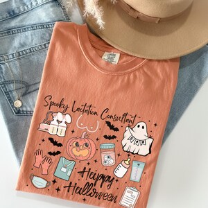 May include: An orange t-shirt with the text "Spooky Lactation Consultant" and "Happy Halloween" with Halloween-themed graphics. The shirt is paired with light blue jeans and a tan hat.
