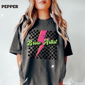 May include: A gray t-shirt with a black and white checkered pattern, a pink lightning bolt, and the text "Brow Artist" in green letters.