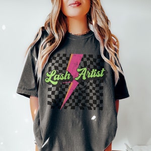 May include: A black t-shirt with a checkered pattern and a pink lightning bolt graphic. The text "Lash Artist" is written in green cursive letters on the shirt.