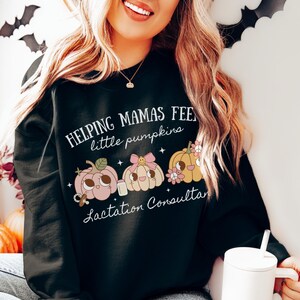 May include: Black sweatshirt with the text "Helping Mamas Feed Little Pumpkins Lactation Consultant." The design features three cartoon pumpkins in pink, peach, and yellow with pacifiers, bows, and flowers. The sweatshirt is worn by a person.