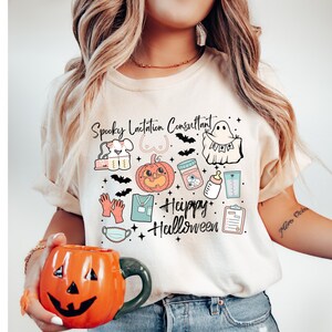 May include: A beige t-shirt with a Halloween-themed graphic that includes a pumpkin, ghost, and the text "Spooky Lactation Consultant" and "Happy Halloween". The person is holding an orange ceramic jack-o'-lantern mug.
