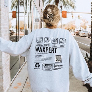 May include: A grey sweatshirt with the text "WAXPERT" and a QR code. The text "TRAINED & KNOWLEDGEABLE", "HYDRATED & DRAMA FREE", "SPF REQUIRED", and "RADIANT & GLOWING" are printed in black boxes on the sweatshirt. The text "@waxperthandle" is printed below the word "WAXPERT".