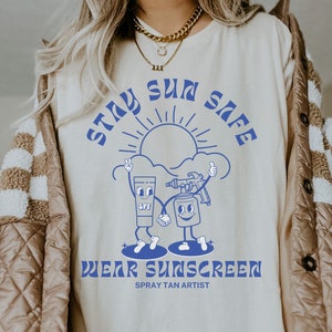 May include: A white t-shirt with blue text that says "Stay Sun Safe" and "Wear Sunscreen" with a sun, a tube of sunscreen, and a spray bottle of sunscreen graphic. The text "Spray Tan Artist" is also printed on the shirt.