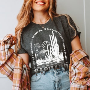 May include: A woman wearing a dark gray t-shirt with a white graphic print of a cactus landscape. The graphic includes the text "Home to America's Largest Cacti & Magnificent Desert Sunsets" and "1994". She is also wearing a plaid shirt and blue jeans.