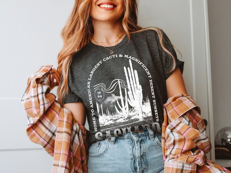 May include: A woman wearing a dark gray t-shirt with a white graphic print of a cactus landscape. The graphic includes the text "Home to America's Largest Cacti & Magnificent Desert Sunsets" and "1994". She is also wearing a plaid shirt and blue jeans.
