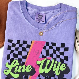 May include: Purple t-shirt with a black and white checkered pattern, a pink lightning bolt, and the text "Line Wife" in green.