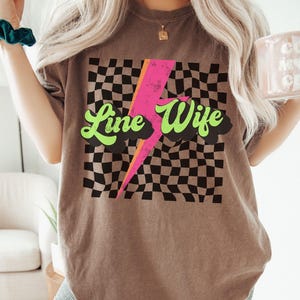 May include: A brown t-shirt with a black and white checkered pattern, a pink lightning bolt, and the text "Line Wife" in green.