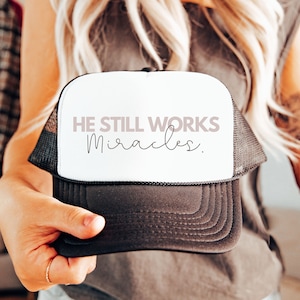 May include: A black and white trucker hat with a white front panel. The front panel has the text "HE STILL WORKS miracles." in pink and black lettering.