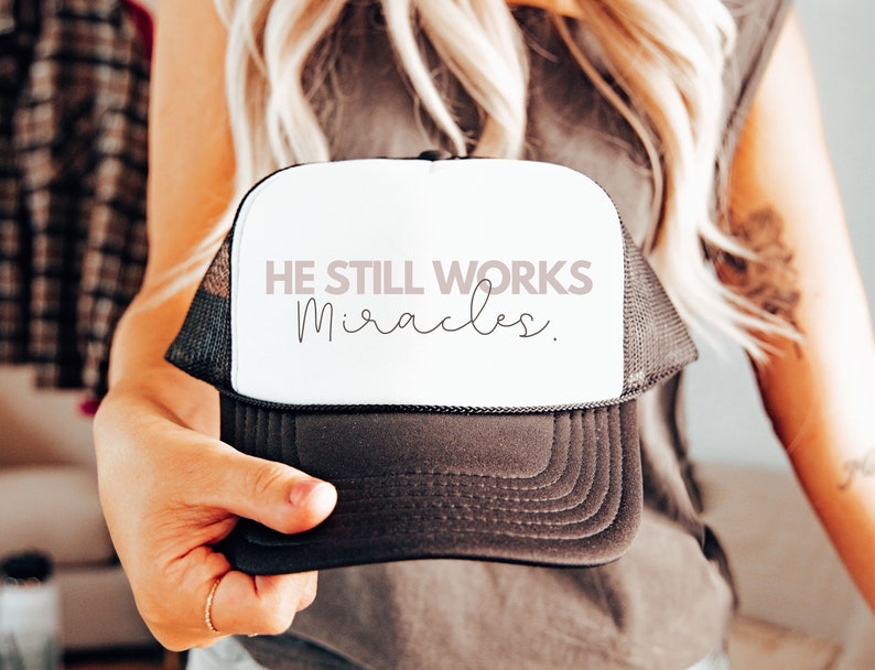 May include: A black and white trucker hat with a white front panel. The front panel has the text "HE STILL WORKS miracles." in pink and black lettering.