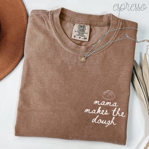 May include: Espresso-colored Comfort Colors t-shirt with the text "mama makes the dough" in white script. A silver necklace with a pendant is draped across the shirt. The shirt is size S-P-CH.