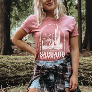 May include: A pink t-shirt with a white graphic design featuring a cactus and the text "Home to America's Largest Cacti & Magnificent Desert Sunsets, Saguaro National Park".