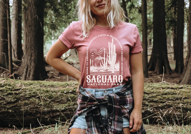 May include: A pink t-shirt with a white graphic design featuring a cactus and the text "Home to America's Largest Cacti & Magnificent Desert Sunsets, Saguaro National Park".