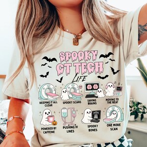 May include: A cream-colored t-shirt featuring a graphic print with the text "SPOOKY CT TECH LIFE" in pink. The design includes various illustrations of ghosts interacting with medical imaging equipment, including CT scanners, monitors, and IV poles.