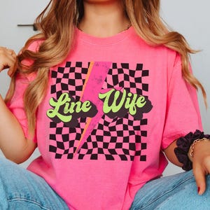May include: Pink t-shirt with a black and white checkered pattern, a lightning bolt, and the text "Line Wife" in a retro font.