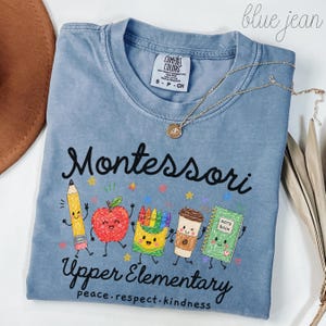 May include: A blue jean colored t-shirt featuring a graphic with the text Montessori Upper Elementary peace respect kindness. The design includes cartoon illustrations of a pencil, apple, crayons, coffee cup, and a notebook with small stars.