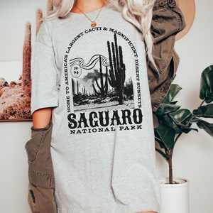 May include: A gray t-shirt with a black graphic design featuring a Saguaro cactus and the text "Home to America's Largest Cacti & Magnificent Desert Sunsets, Saguaro National Park".