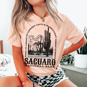 May include: A pink t-shirt with a black graphic design featuring a silhouette of a desert landscape with saguaro cacti and the text "Home to America's Largest Cacti & Magnificent Desert Sunsets" and "Saguaro National Park".