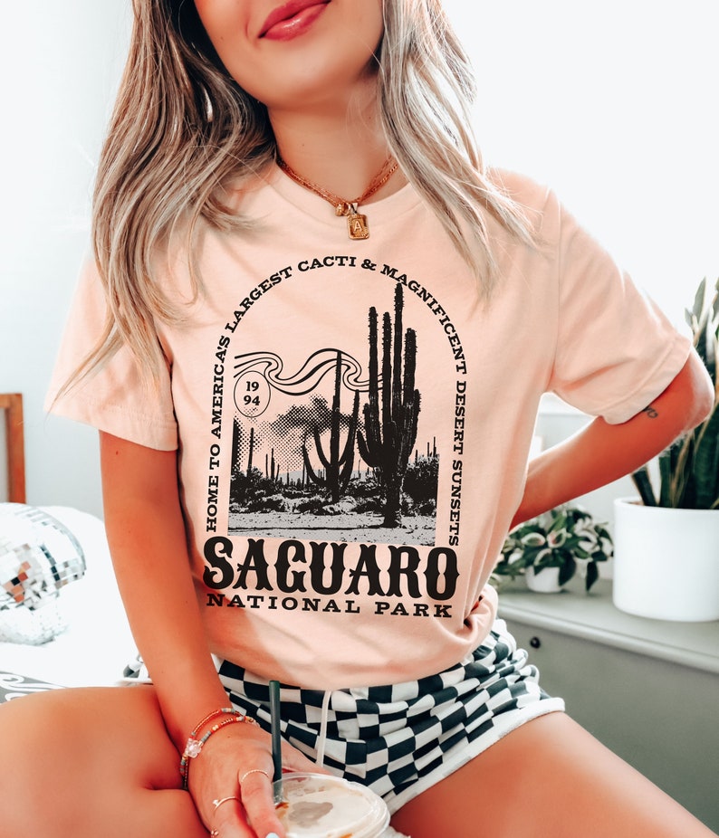 May include: A pink t-shirt with a black graphic design featuring a silhouette of a desert landscape with saguaro cacti and the text "Home to America's Largest Cacti & Magnificent Desert Sunsets" and "Saguaro National Park".