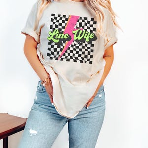 May include: A woman wearing a light beige t-shirt with a black and white checkered pattern, a pink lightning bolt, and the text "Line Wife" in green letters. The shirt is a relaxed fit and has a crew neck.