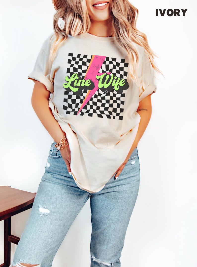 May include: A woman wearing a light beige t-shirt with a black and white checkered pattern, a pink lightning bolt, and the text "Line Wife" in green letters. The shirt is a relaxed fit and has a crew neck.