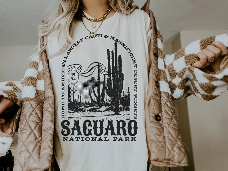 May include: A white t-shirt with a black graphic design featuring a desert landscape with cacti and the text "Home to America's Largest Cacti & Magnificent Desert Sunsets Saguaro National Park".