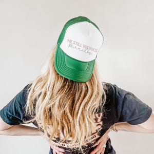 May include: A green and white trucker hat with the text "He Still Works Miracles" in white and pink lettering.