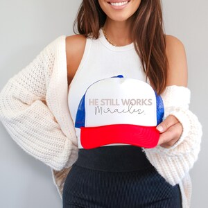 May include: White trucker hat with a red brim and blue mesh back. The hat has the text "He Still Works Miracles." in black and pink lettering.