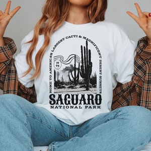 May include: White t-shirt with a black graphic design featuring a silhouette of a desert landscape with cacti and the text "Home to America's Largest Cacti & Magnificent Desert Sunsets Saguaro National Park".