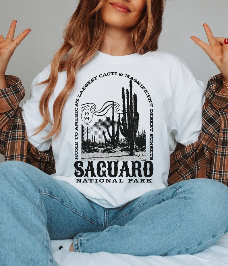 May include: White t-shirt with a black graphic design featuring a silhouette of a desert landscape with cacti and the text "Home to America's Largest Cacti & Magnificent Desert Sunsets Saguaro National Park".