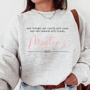 May include: A light gray sweatshirt with the text "She turned her cants into cans and her dreams into plans, Master's 2023" in black and pink script. The sweatshirt has long sleeves and a crew neck.