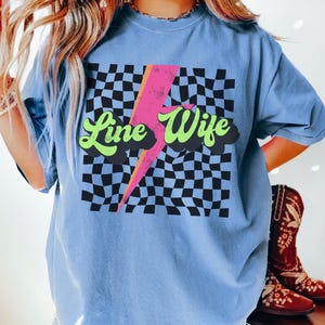 May include: A light blue t-shirt with a checkered pattern and a lightning bolt graphic. The text "Line Wife" is written in a retro font.