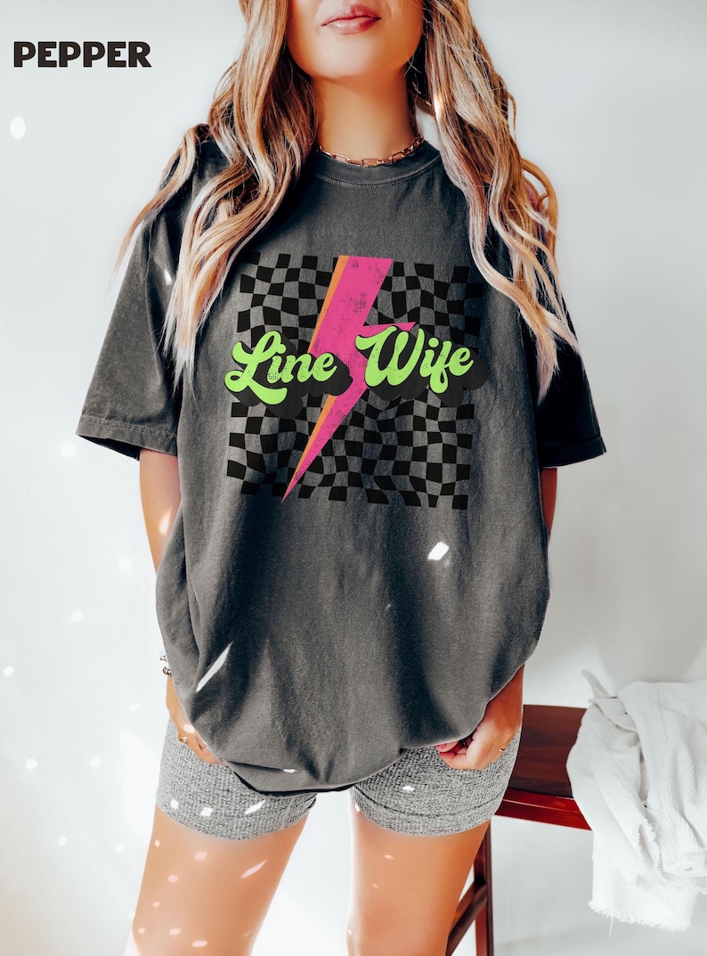 May include: A gray t-shirt with a checkered pattern and a pink lightning bolt graphic. The text "Line Wife" is written in green and pink letters.
