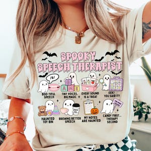 May include: A cream-colored t-shirt featuring a graphic with the text SPOOKY SPEECH THERAPIST in pink, surrounded by illustrations of ghosts, bats, and speech therapy items with captions like BOO-TIFUL SPEECH and MY NOTES ARE HAUNTED.