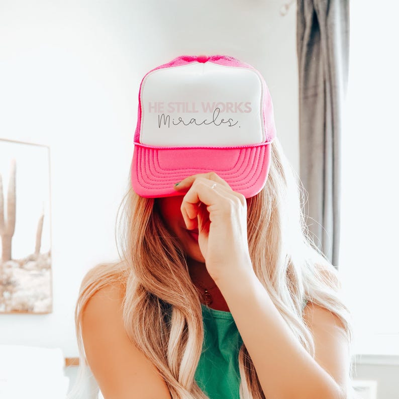 May include: Pink and white trucker hat with the text "He Still Works Miracles." in white lettering.