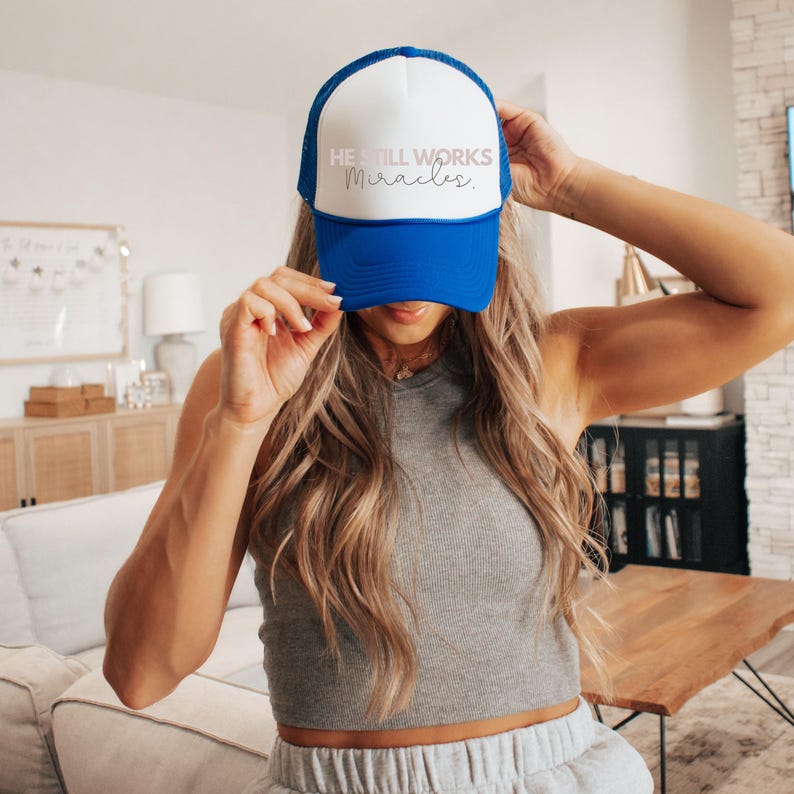 May include: A blue and white trucker hat with the text "He Still Works Miracles" in pink and white lettering.