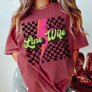 May include: A brick red t-shirt with a black and white checkered pattern, a pink lightning bolt, and the text "Line Wife" in green and black.