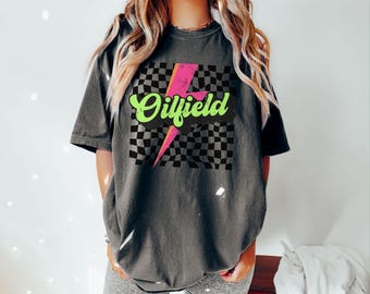 Puede incluir: Una camiseta negra con un patrón a cuadros y un gráfico de rayo rosa y verde. El texto "Oilfield" está escrito en una fuente retro.