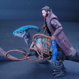 Pu&ograve; includere: Un giocattolo di dinosauro blu e marrone con una sella e una corda marrone. Una action figure di un uomo &egrave; a cavallo del dinosauro.