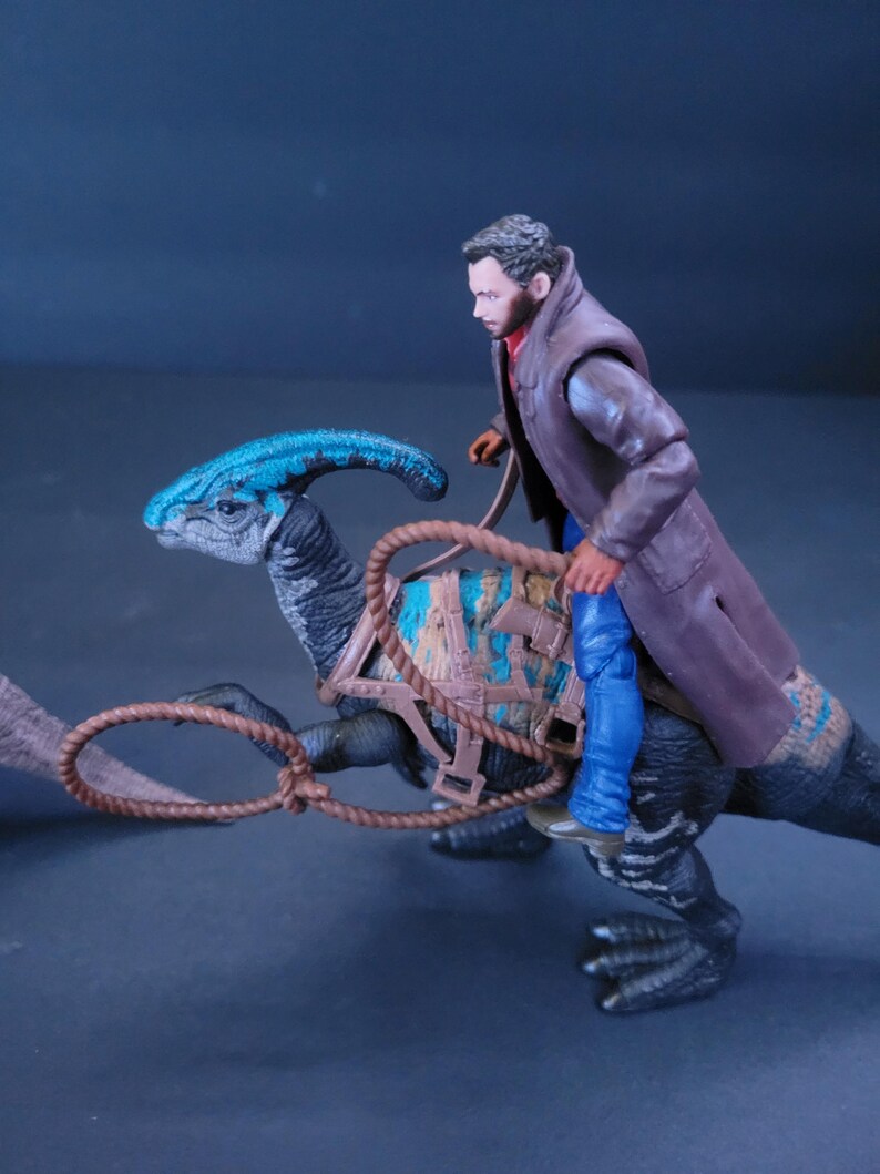 Pu&ograve; includere: Un giocattolo di dinosauro blu e marrone con una sella e una corda marrone. Una action figure di un uomo &egrave; a cavallo del dinosauro.