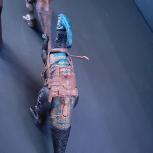 Pu&ograve; includere: Un giocattolo di dinosauro nero e blu con un'imbracatura marrone. Il dinosauro ha una cresta blu sulla testa e una lunga coda.