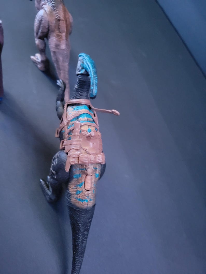 Pu&ograve; includere: Un giocattolo di dinosauro nero e blu con un'imbracatura marrone. Il dinosauro ha una cresta blu sulla testa e una lunga coda.