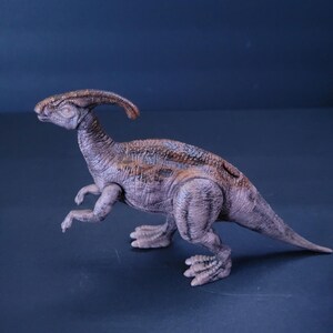 Pu&ograve; includere: Un giocattolo di dinosauro marrone e grigio con un collo lungo e una cresta sulla testa. Il dinosauro &egrave; in piedi su una superficie blu.