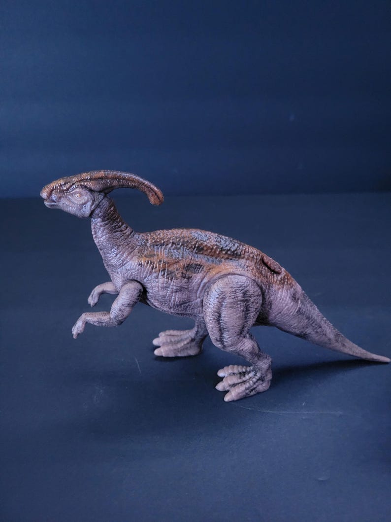 Pu&ograve; includere: Un giocattolo di dinosauro marrone e grigio con un collo lungo e una cresta sulla testa. Il dinosauro &egrave; in piedi su una superficie blu.