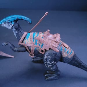 Pu&ograve; includere: Un giocattolo di dinosauro nero e blu con una sella e redini marroni. Il dinosauro ha un collo lungo e una cresta sulla testa.