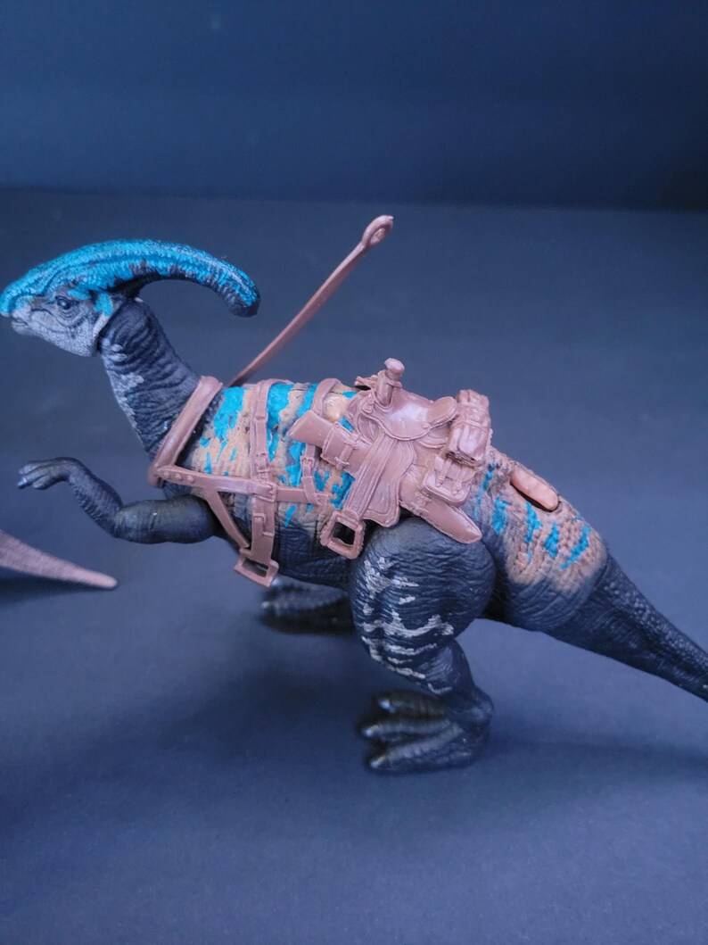 Pu&ograve; includere: Un giocattolo di dinosauro nero e blu con una sella e redini marroni. Il dinosauro ha un collo lungo e una cresta sulla testa.
