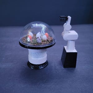 Peut inclure: Un bras robotique blanc avec une main noire, à côté d'un dôme en verre transparent avec une petite base de mousse verte et plusieurs petits œufs mouchetés à l'intérieur.