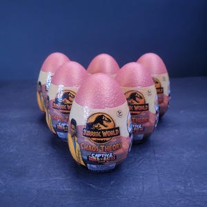 Puede incluir: Seis cápsulas de juguetes rosas en forma de huevo de "Chaos Theory" de Jurassic World. Cada cápsula tiene una etiqueta con el logotipo de Jurassic World, el texto "Chaos Theory" y el logotipo de Captivz Build & Battle. Las cápsulas están dispuestas en forma de pirámide.