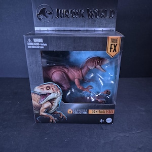 Puede incluir: Figura de acción Ornitholestes de la colección Hammond de Jurassic World en caja. La figura es un dinosaurio marrón rojizo con detalles. El embalaje incluye el logotipo de Jurassic World y una advertencia.