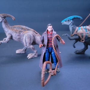 Pu&ograve; includere: Una action figure di un uomo che indossa una giacca marrone, una camicia a righe rosse e bianche, jeans blu e tiene una corda marrone. La figura &egrave; in piedi davanti a due action figure di dinosauri, una marrone e una blu.